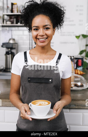 Junge lächelnde Kellnerin halten einer Tasse Latte, stehend an der Bar Schreibtisch im Coffeeshop Stockfoto