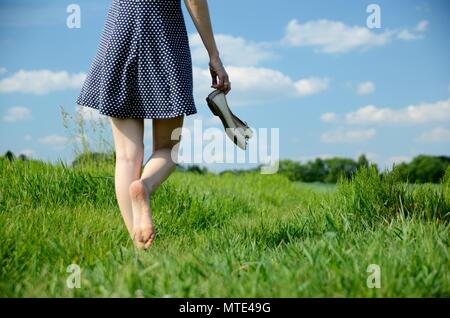 Weibliche Wege auf dem Gras mit barfuß. Junge Frau genießt Spaziergang im Freien, Natur, grüne Wiesen und blauer Himmel. Stockfoto