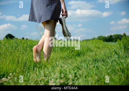 Weibliche Wege auf dem Gras mit barfuß. Junge Frau genießt Spaziergang im Freien, Natur, grüne Wiesen und blauer Himmel. Stockfoto