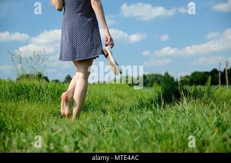 Weibliche Wege auf dem Gras mit barfuß. Junge Frau genießt Spaziergang im Freien, Natur, grüne Wiesen und blauer Himmel. Stockfoto