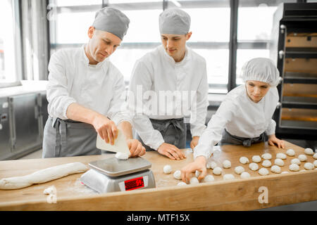 Drei junge Bäcker in Uniform bilden Teig zum backen auf dem hölzernen Tisch zusammen, der auf die moderne Fertigung Stockfoto