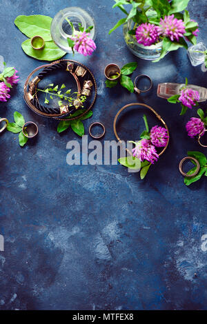 Goldene Armbänder und Ringe in einem femininen Stil Konzept mit Klee Blumen auf einem dunklen Hintergrund. Schönen Sommer Zubehör. Stockfoto