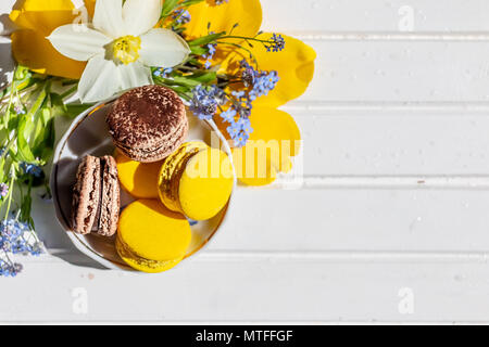 Macarons oder makronen Dessert süße schön zu essen. Schokolade und Zitrone Desserts auf einem weißen Holztisch. leckeren Nachtisch flach und Narzissen und Vergißmeinnicht Blumen. Stockfoto