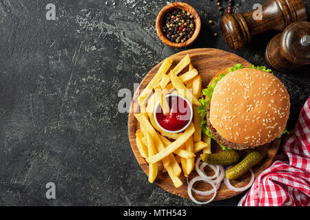 Burger und Pommes frites auf Holzbrett auf dunklem Stein Hintergrund. Hausgemachten Burger oder Cheeseburger, Pommes frites und Ketchup. Leckeres Sandwich. Ansicht von oben mit Co Stockfoto