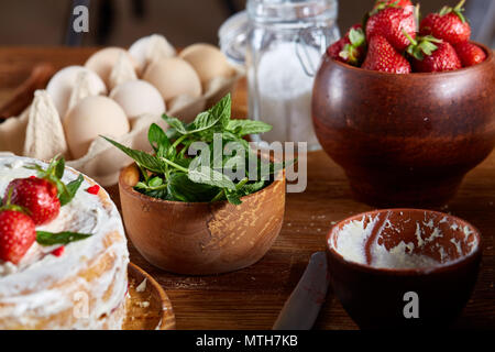 Backen Hintergrund mit Erdbeerkuchen mit Schlagsahne Sauerrahm durch organische Inhaltsstoffe über Holztisch umgeben, selektiven Fokus. Hausgemachte Stockfoto