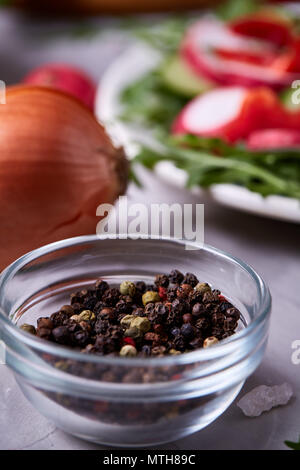 Glasschale mit getrockneten Piment Samen vor der Platte mit frischem Gemüse Salat auf weiße strukturierte Hintergrund, selektiver Fokus, senkrecht. Nesessary s Stockfoto