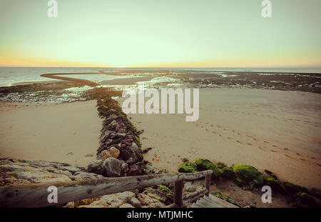 Holzgeländer die zum Strand von Sanlúcar de Barrameda mit grünen Steinen am Ufer und den Sonnenuntergang im Hintergrund Stockfoto