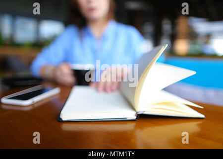 Close up Notebook in der Nähe von Smartphone mit Mädchen im Cafe im Hintergrund, verschwommenes Foto. Stockfoto