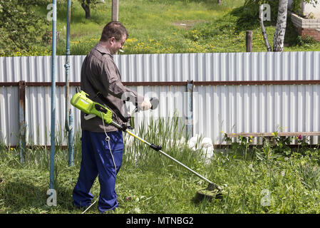 Ein Mann in Gläser mit einem trimer mäht das Gras im Hof eines Hauses in dem Dorf, bringt Ordnung in das Gebiet des Landes Stockfoto