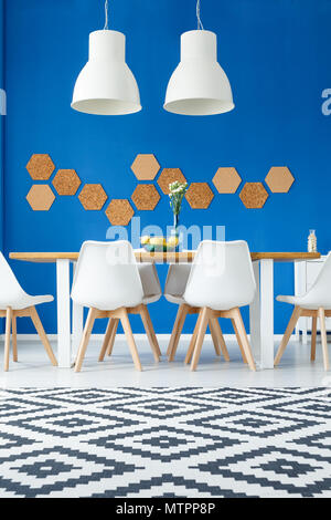 DIY Wabe Cork auf blauen Wand in der modernen natürlichen Esszimmer mit hölzernen Gemeinschaftstisch, weißen Stühlen und gemustertem Teppich Stockfoto