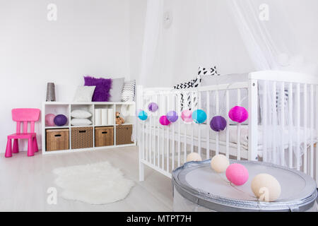 Modern gestaltetes Interieur Baby's Schlafzimmer mit weißen Wiege mit bunten Kugeln geschmückt. An der gegenüberliegenden Wand Regal mit Boxen und Kissen Stockfoto