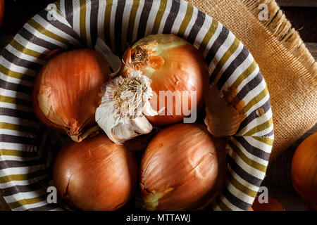 Noch immer leben von Zwiebeln und Knoblauch Kopf in einem Weidenkorb auf einem Sack und Holzplatten. Von oben gesehen. Im rustikalen Stil. Bild horizontal. Stockfoto