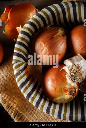 Noch immer leben von Zwiebeln und Knoblauch Kopf in einem Weidenkorb auf einem Sack und Holzplatten. Von oben gesehen. Im rustikalen Stil. Bild vertikal. Stockfoto