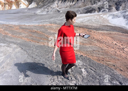 Frau in einem Business Red Suit, Festgefahren im Schlamm in der Mitte eines wilden Canyon, schaut auf das Tablett in der Hand. Stockfoto
