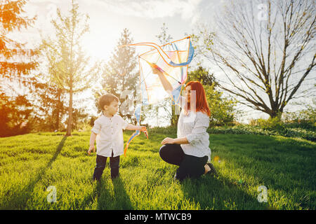 Schöne und stilvolle rothaarige Mama in eine weiße Bluse spielen mit ihren kleinen süßen Sohn in einem Sommer sonnigen Park mit einem Kite Stockfoto