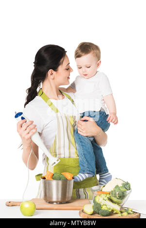 Lächelnde Mutter mit Sohn und Stabmixer bei Tisch mit Apfel und Gemüse auf weißem Hintergrund Stockfoto