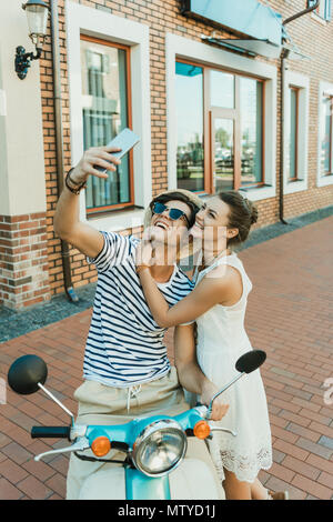 Lächelnde junge Paar unter selfie auf dem Smartphone beim Sitzen auf Roller im Freien Stockfoto