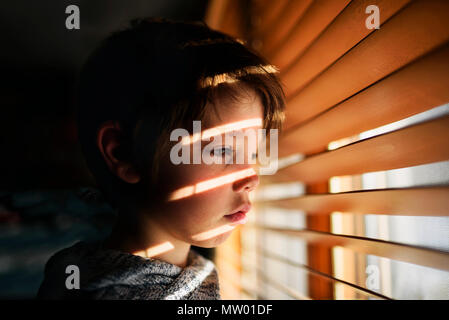 Junge durch ein Fenster schauen durch Jalousien Stockfoto