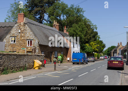 Die Hauptstraße durch das Dorf wenig Harrowden, mit einem schönen reetgedeckten Gebäude auf der linken Seite plus ein einsamer Jogger; Northamptonshire, Großbritannien Stockfoto