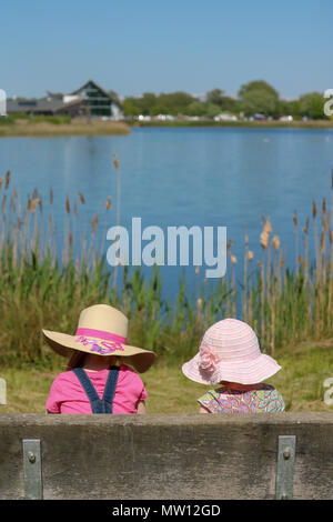 Ein junges Mädchen und ein Kleinkind, Rücken zur Kamera und sowohl in sunhats, sitzen auf einer Bank und die Aussicht über den See an einem herrlich sonnigen Tag an Stanwick Lakes, Northampton, UK betrachten Stockfoto