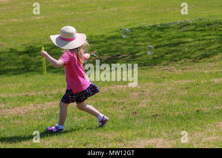 Ein junges Mädchen in hellen Sommer Kleidung und einen Sonnenhut, von der Kamera entfernt, läuft durch einen Park streaming Blasen hinter ihr aus einem bubble Wand an einem sonnigen Sommertag. Stockfoto