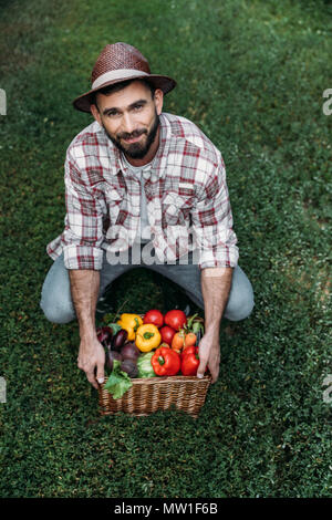 Bärtige Landwirt in Hut holding Korb mit reifem Obst und lächelnd an Kamera Stockfoto