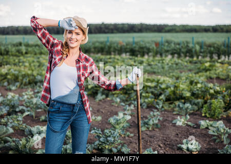 Schöne junge Frau Bauer in Hut holding Hacke und Arbeiten auf dem Feld Stockfoto