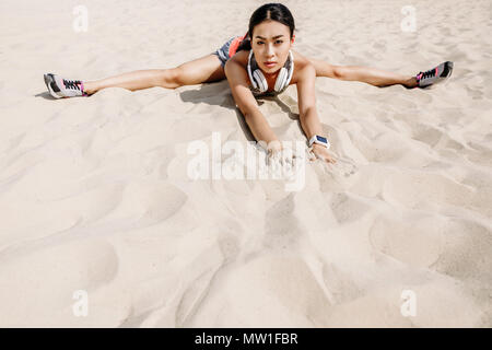 Asiatische Sportlerin mit Kopfhörern tun Split auf Sand Stockfoto
