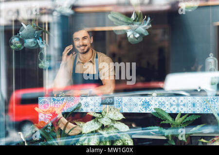 Hübscher junger Floristen sprechen auf Smartphone und lächelnd in die Kamera durch das Fenster in Flower Shop Stockfoto