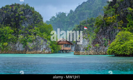 Bambushütte zwischen einigen Felsen unter Regen in der Bucht mit indonesischen Flagge, Pianemo Inseln, Raja Ampat, West Papua, Indonesien Stockfoto