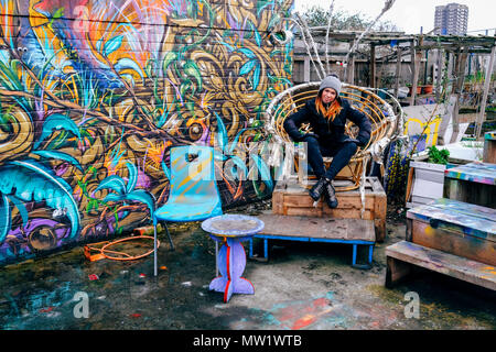 Eine Frau sitzt auf dem ovalen Stuhl neben der Graffiti wall die nomadische Gemeinschaft Garten aus Brick Lane in Shoreditch, East London, England, UK. Stockfoto