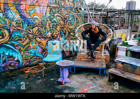 Eine Frau sitzt auf dem ovalen Stuhl neben der Graffiti wall die nomadische Gemeinschaft Garten aus Brick Lane in Shoreditch, East London, England, UK. Stockfoto