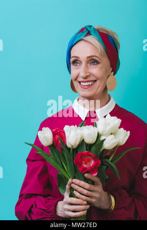 Attraktives Lächeln ältere Frau mit Blumenstrauß aus Frühlingsblumen, isoliert auf Türkis Stockfoto