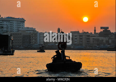 Touristen an Bord eines Abra, einem traditionellen Holz- Fähre, segeln in Richtung der Küste in Dubai Creek bei Sonnenuntergang. Die Skyline der Stadt, Silhouetten, Orange Sky Stockfoto