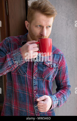 Super Kerl mit Ingwer Haar sein Getränk in rot Tasse genießen. Gut aussehenden Mann tragen Flanellhemd, Schluck Kaffee aus seiner Tasse in suchen Stockfoto