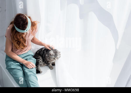 Hohe Betrachtungswinkel und der jungen Frau petting adorable tabby Katze sitzend auf Fensterbank Stockfoto