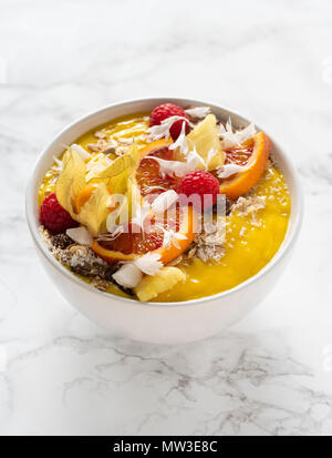 Gelbe mango Smoothie Schüssel mit Müsli, Beeren, Obst dekoriert, Kokos Flocken auf weißem Marmor Hintergrund Stockfoto