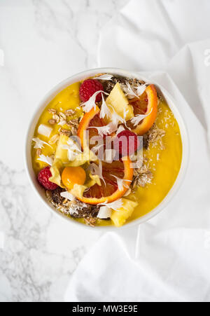 Gelbe mango Smoothie Schüssel mit Müsli, Beeren, Obst dekoriert, Kokos Flocken auf weißem Marmor Hintergrund Stockfoto