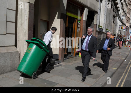 Ein Arbeitnehmer zieht eine grüne Mülltonne Vergangenheit geeignet Geschäftsleute in der Stadt London, der Bezirk aka der Hauptstadt der Square Mile, am 17. Mai 2018 in London, Großbritannien. Stockfoto