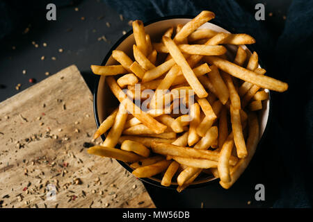 High Angle Shot einige leckere Pommes frites in eine weiße keramische Schüssel serviert, auf einem dunkelgrauen rustikalen Holztisch gelegt Stockfoto