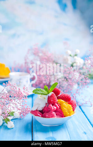 Eis von hellen Blüten mit Himbeeren und Minze in einer Platte, auf blauem Holz- Hintergrund. Eine Tasse Kaffee am Morgen und Licht und Blumen. Vintage Foto. Platz kopieren Stockfoto