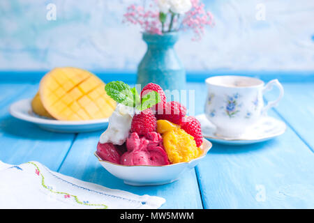 Eis von hellen Farben mit Mango, Himbeeren und Minze in einer Platte, auf blauem Holz- Hintergrund. Eine Tasse Kaffee am Morgen und Licht und Blumen. Vintage Foto. Platz kopieren Stockfoto
