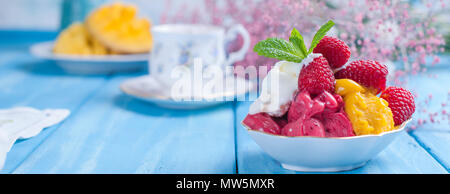 Eis von hellen Farben mit Mango, Himbeeren und Minze in einer Platte, auf blauem Holz- Hintergrund. Eine Tasse Kaffee am Morgen und Licht und Blumen. Vintage Foto. Banner. Platz kopieren Stockfoto