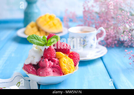 Eis von hellen Farben mit Mango, Himbeeren und Minze in einer Platte, auf blauem Holz- Hintergrund. Eine Tasse Kaffee am Morgen und Licht und Blumen. Vintage Foto. Banner. Platz kopieren Stockfoto