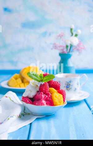Eis von hellen Blüten mit Himbeeren und Minze in einer Platte, auf blauem Holz- Hintergrund. Eine Tasse Kaffee am Morgen und Licht und Blumen. Vintage Foto. Platz kopieren Stockfoto