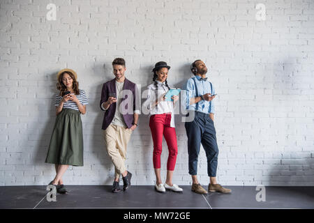 Junge stilvolle Freunde mit digitalen Geräten beim Stehen in der Nähe von Mauer Stockfoto