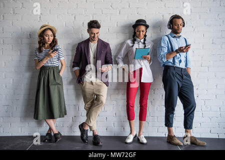 Junge stilvolle Freunde mit digitalen Geräten beim Stehen in der Nähe von Mauer Stockfoto