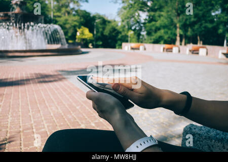 Junge Frau wischen smart phone Screen mit antibakterieller Stoff, beim Sitzen im Park in der Nähe von Brunnen. Weibliche Hände halten mobile Gerät und reinigen Stockfoto