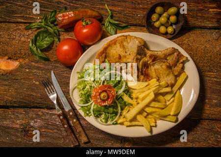 Oben Ansicht von Medium gegrillte Lampe mit Kartoffeln französischen auf weißem Teller serviert, Tomaten, Karotten, Messer und Gabel auf Schneidebrett Chop, auf hölzernen Hintergrund Stockfoto