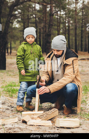 Hübscher junger Vater und Sohn Schneiden von Holz für Lagerfeuer im Wald Stockfoto